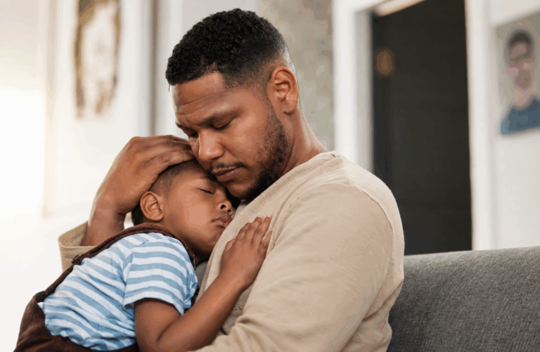 Father comforting his sleeping son while sitting together on a couch at home.